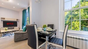 Ett modernt vardagsrum med en platt-TV som visar "NETFLIX", en matplats med ett bord dukat för två personer och ett stort fönster med utsikt över träd.