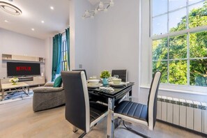 A modern living room with a flat-screen TV displaying 'NETFLIX', a dining area with a table set for two, and a large window offering a view of trees.