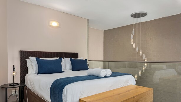 Ett modernt hotellrum med en stor säng, en träbänk, ett sängbord med en lampa och en vägg av glas som avskärmning.