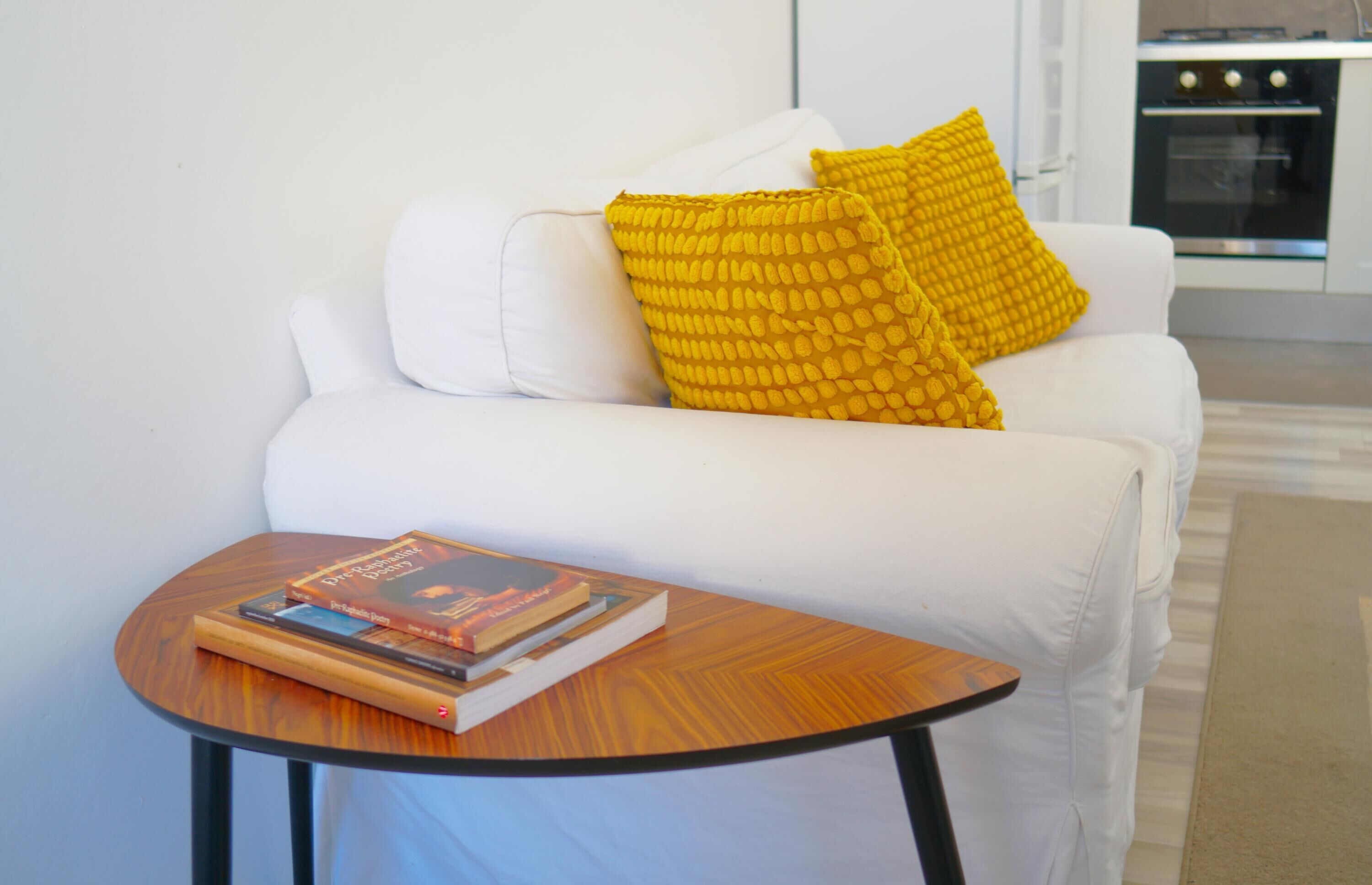 A white sofa with two yellow pillows, a small wooden table with books, and a kitchen in the background.
