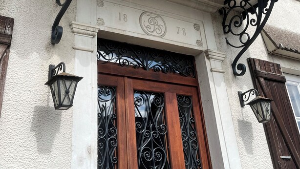Fachada de edificio con un balcón decorativo de hierro forjado, una puerta de madera con detalles de hierro y dos faroles fijados a la pared.