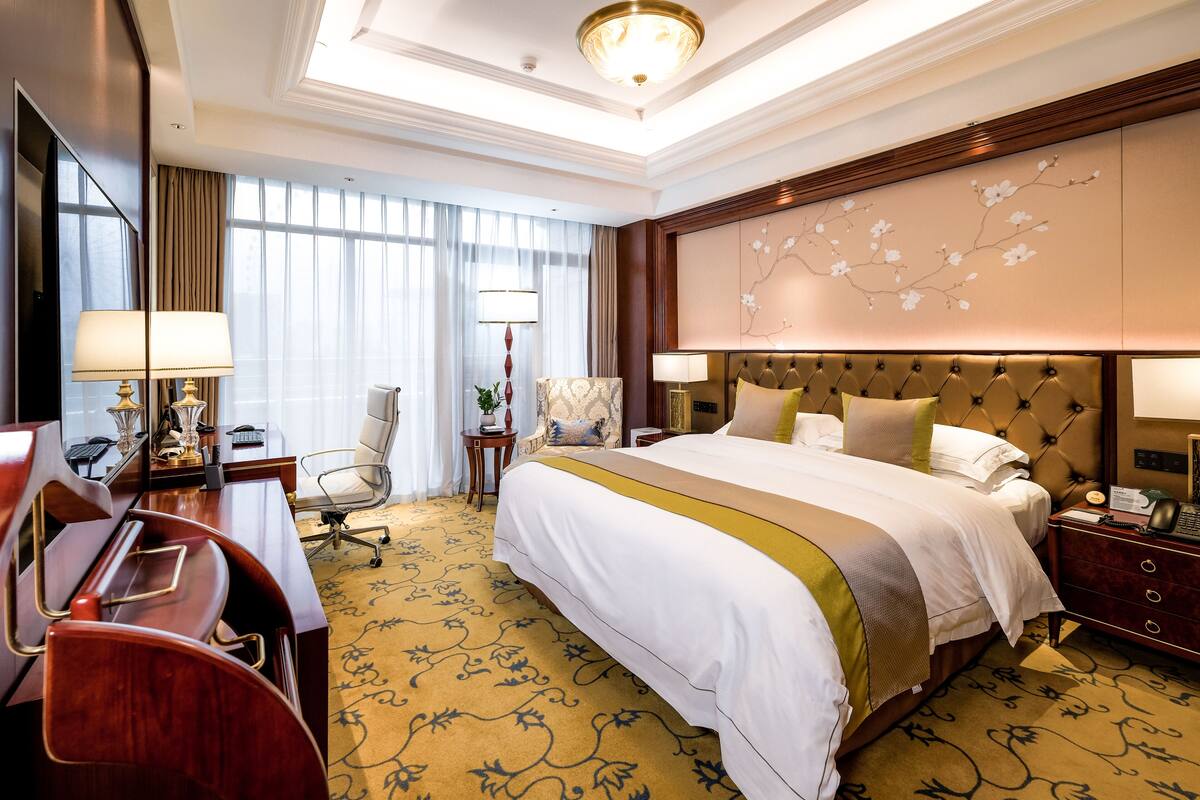 Quarto de hotel com uma cama grande, uma escrivaninha, uma cadeira, um abajur e um detalhe decorativo na parede.