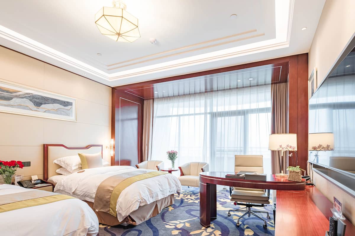 Quarto de hotel com duas camas, uma escrivaninha, cadeiras, uma janela grande com cortinas e um candelabro.