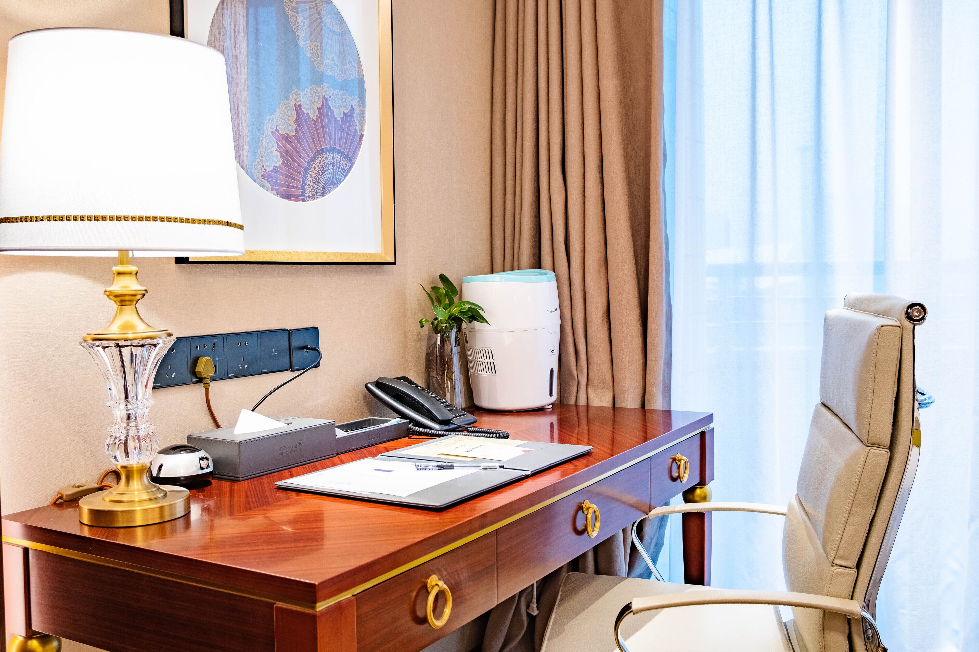 Ein Hotelzimmer mit einem Holztisch, einer Lampe, einem Telefon, einem Drucker, einer Pflanze und einem Stuhl.