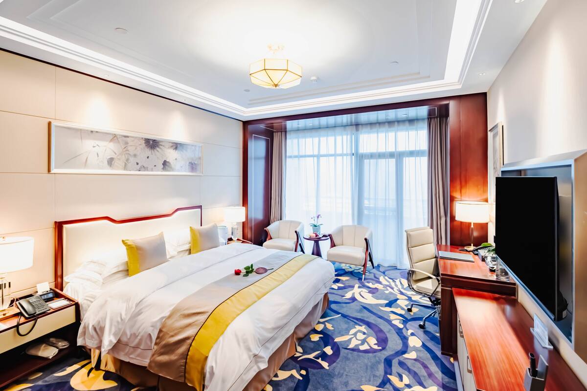 Quarto de hotel com uma cama grande, uma escrivaninha com televisão de tela plana, duas cadeiras, uma mesinha de cabeceira com abajur e um janelão com cortinas.