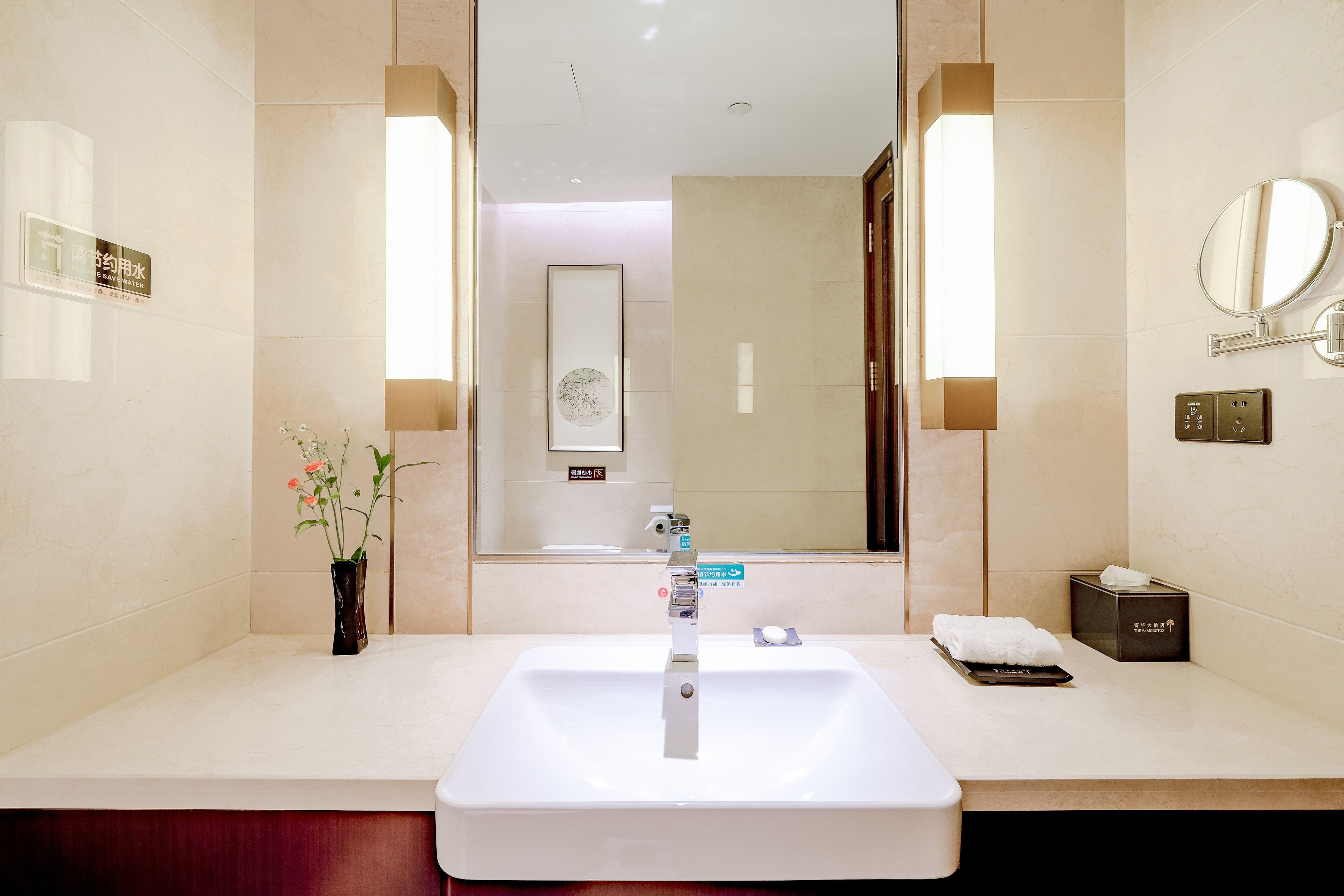 Une salle de bain d’hôtel avec un grand miroir, un lavabo, un vase de fleurs et une boîte de mouchoirs.