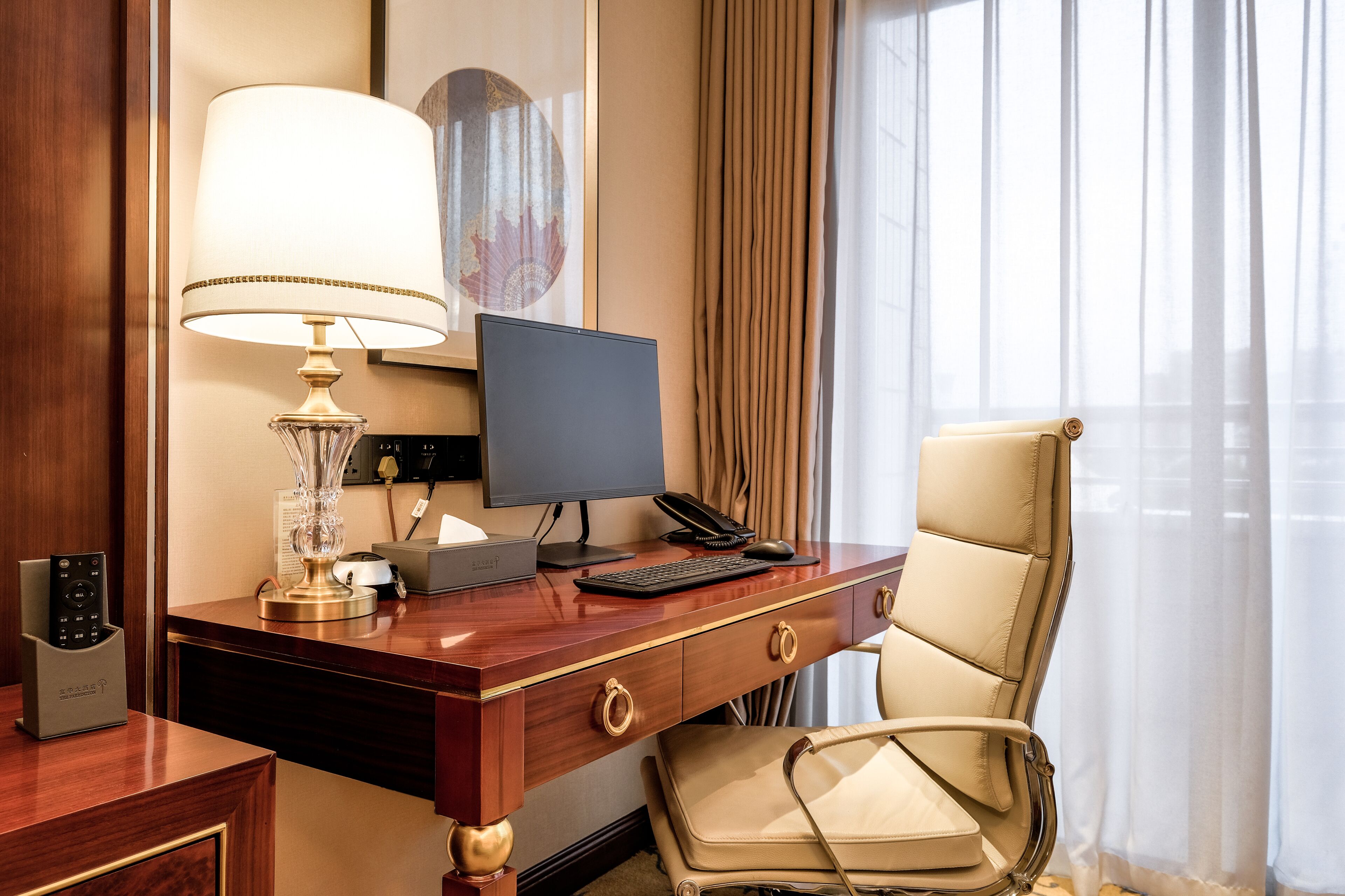 A hotel room with a wooden desk, a computer, a lamp, a chair, and a window with curtains.