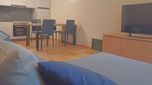 A compact living space with a kitchenette, a dining area with chairs, a television on a wooden cabinet, and a bed with blue bedding. - Acquaseria 310 in Ponte di Legno (Ponte di Legno)