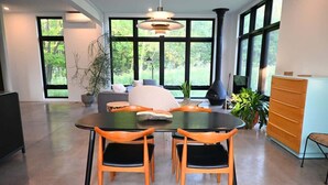 Et moderne spiseområde med et rundt bord og stole, en sofa og et stort vindue med udsigt til grønne områder.