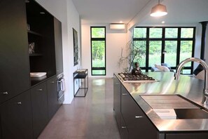 A modern kitchen with dark cabinetry, a stainless steel island, and large windows offering a view of greenery.