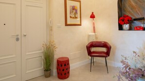 Un couloir avec une porte blanche, un tableau encadré au mur, une chaise rouge, une lampe rouge et une plante en pot.