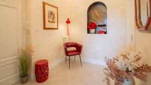 Un couloir avec une chaise rouge, une plante en pot, un tableau encadré et une fenêtre ornée de fleurs.