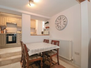 A dining area with a table and chairs, a kitchen in the background, and a wall clock. - Scrambles Cottage (Stoke-sub-Hamdon)