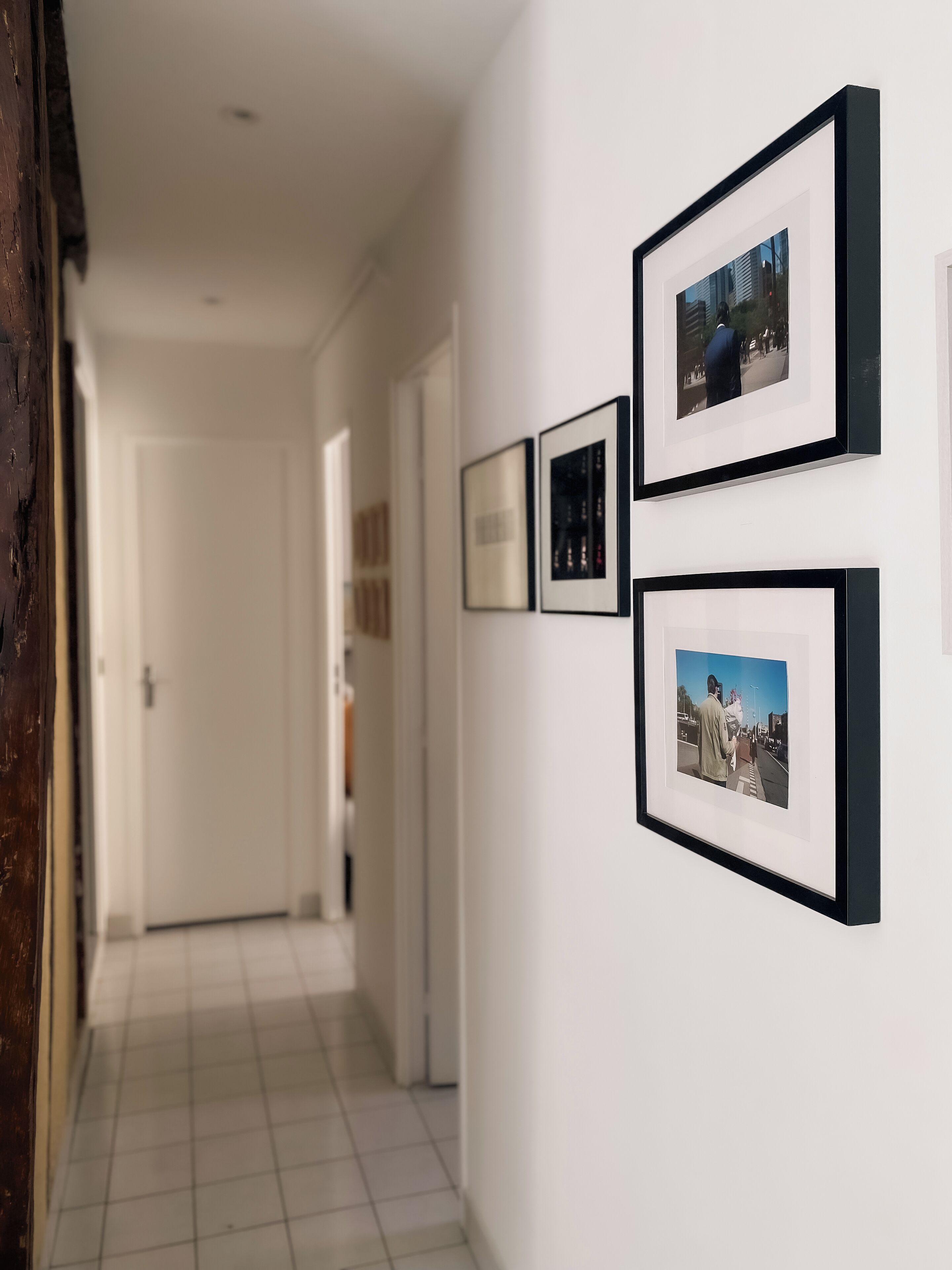 A hallway with framed photographs on the wall, a door at the end, and a tiled floor.