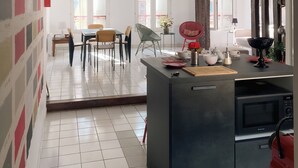 A modern kitchen with a black countertop, a red kettle, and a microwave. There is a dining area with chairs and a table, and a hallway with a tiled floor and a wall with a color pattern.