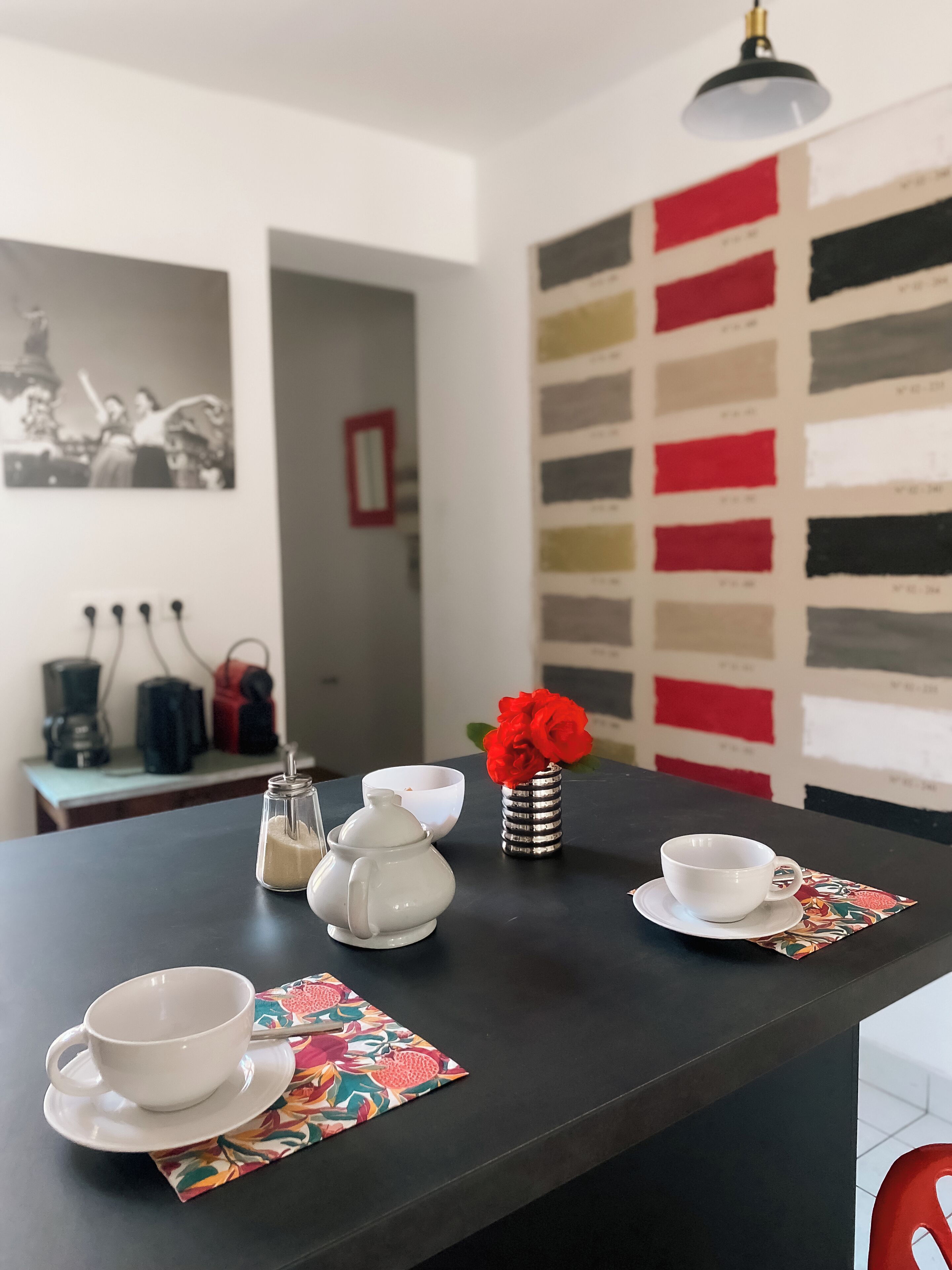 Un tavolo da pranzo con una tazza bianca, un piattino e un piccolo vaso con fiori rossi. Sul tavolo ci sono due teiere bianche e una bottiglia. La parete sullo sfondo presenta un motivo con una palette di colori.