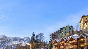 A mountain resort with wooden chalets, a green building with scaffolding, and snow-covered mountains in the background. - Villa Nice View in Pontagna (Temu)