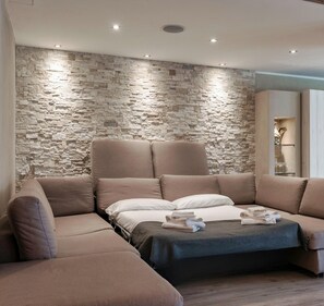 A modern living room with a stone wall, a sectional sofa, and a bed with a dark throw blanket. - La Tavolata in Incudine (Incudine)
