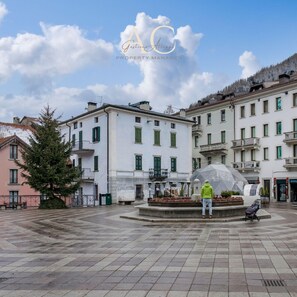 A public square with a fountain, surrounded by multi-story buildings with balconies and green shutters. - Square Apartment in Ponte di Legno (Ponte di Legno)