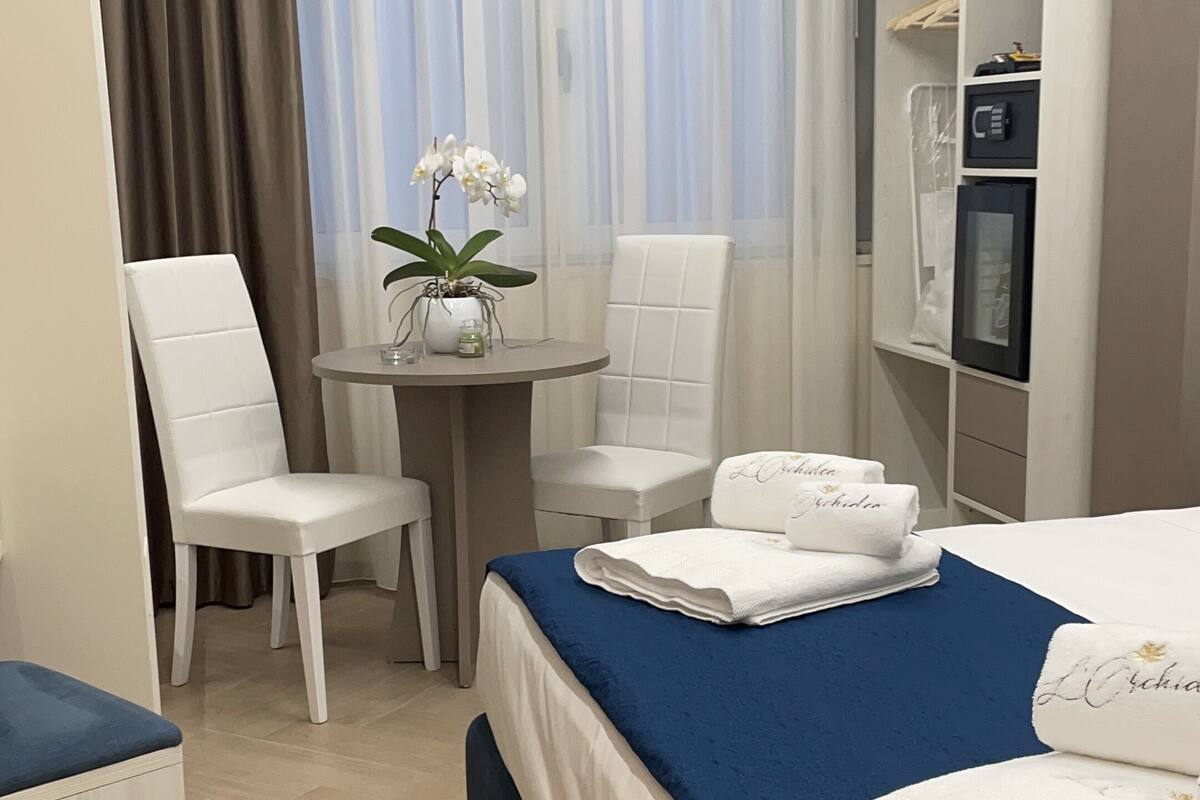 A hotel room with a bed, two white chairs, a small table with a vase of flowers, a wardrobe, and a desk.