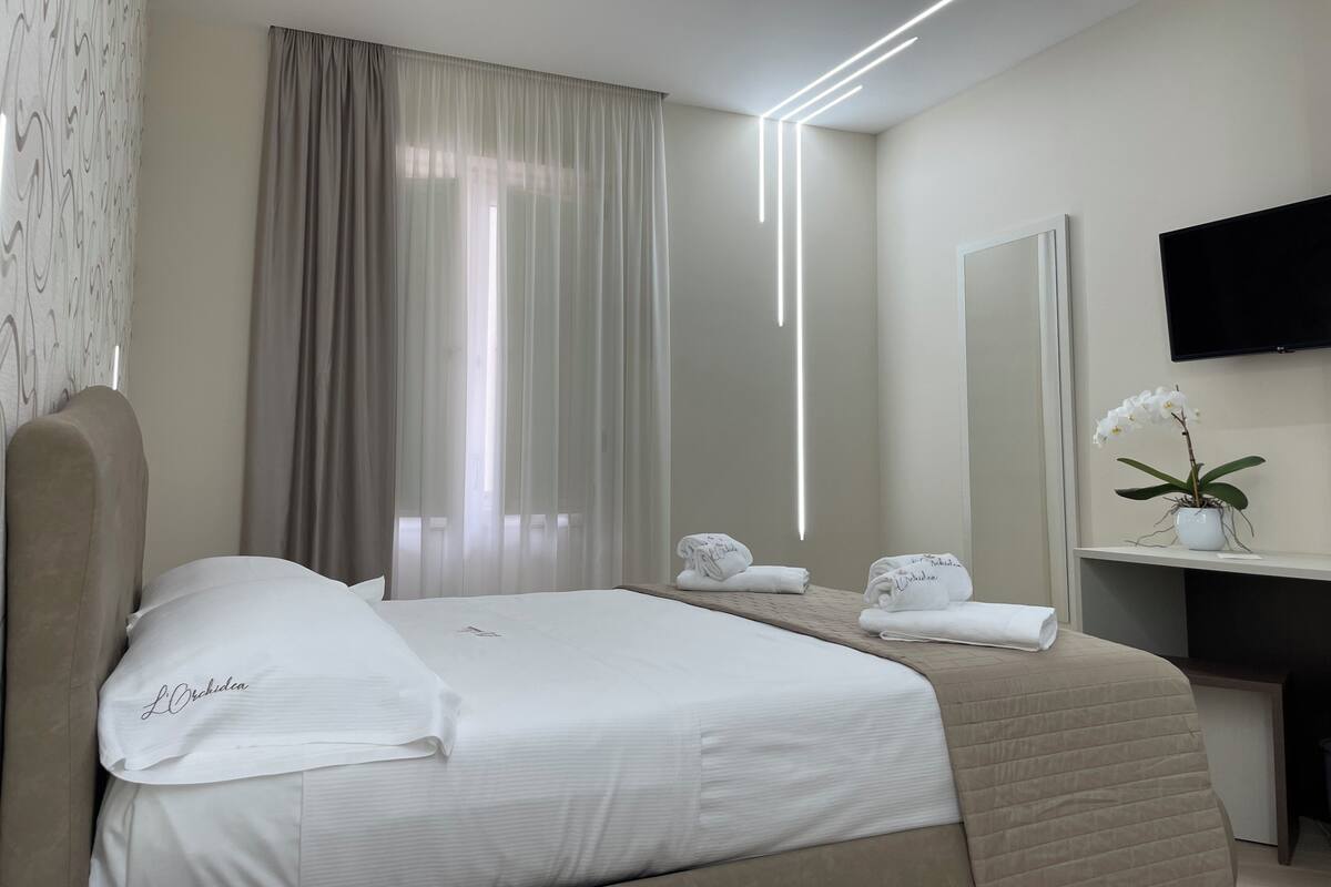 A modern hotel room with a neatly made bed, a large window with curtains, a flat-screen TV, a small desk with a potted orchid, and a mirror.