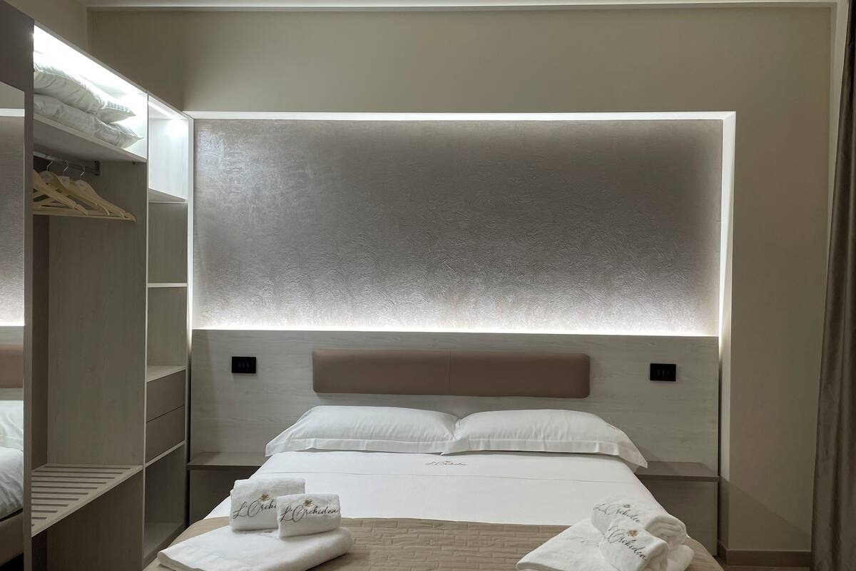 A hotel room with a large bed, a wardrobe, and a headboard with built-in lighting.