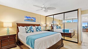 A hotel room with a large bed, a nightstand, a lamp, a ceiling fan, a painting, and a sliding glass door leading to a balcony. - Beachfront bliss near the Heart of Kaanapali shopping (Lahaina)