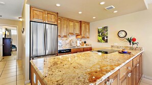 A kitchen with wooden cabinets, a stainless steel refrigerator, granite countertops, and a clock on the wall. - Beachfront bliss near the Heart of Kaanapali shopping (Lahaina)