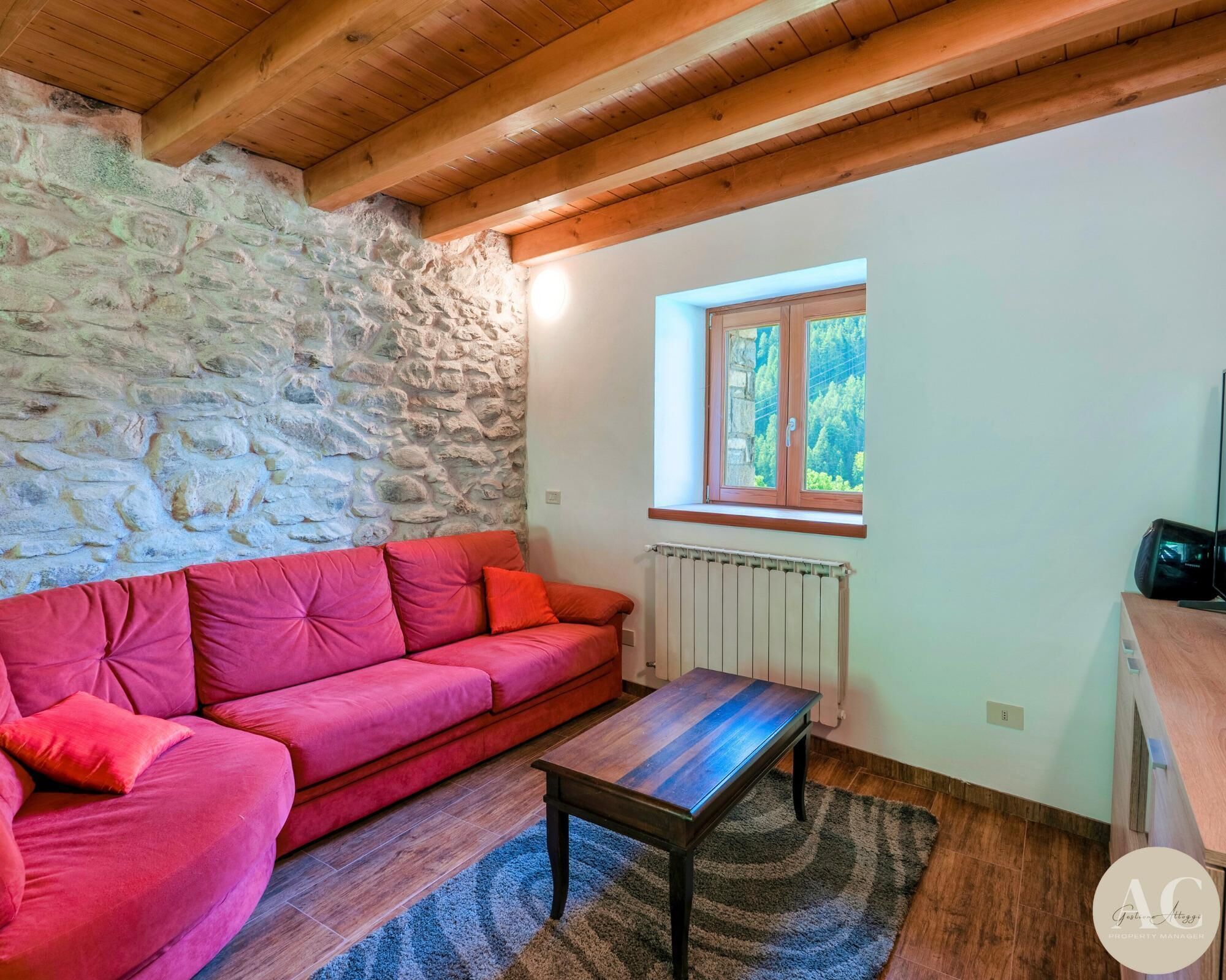 Una habitación acogedora con pared de piedra, un sofá rojo, una mesa de madera y una ventana con vistas al verdor.