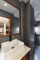 Un baño moderno con un lavamanos blanco rectangular, paredes de baldosas gris oscuro y un espejo con marco de madera.