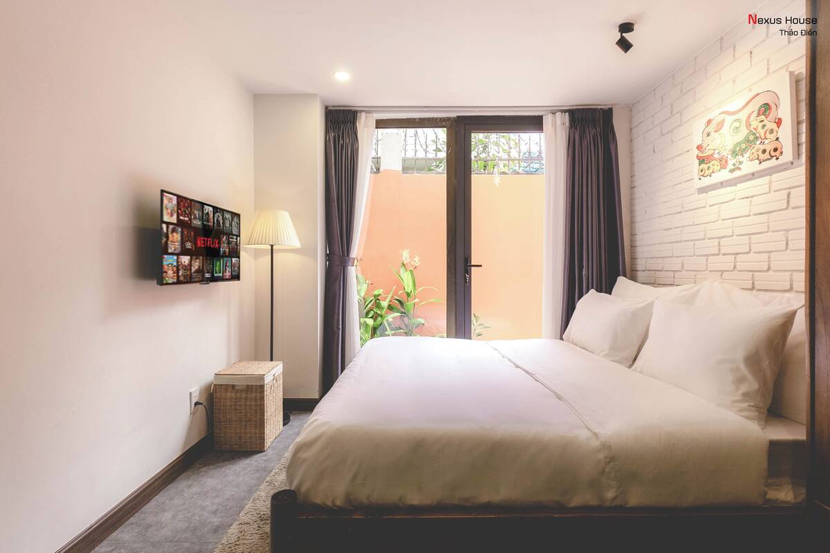 A bedroom with a bed, a wall-mounted TV, a lamp, and a view of the outside.