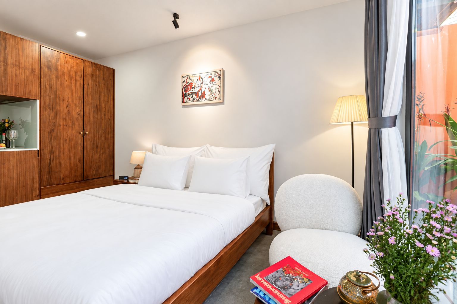 A hotel room with a large bed, a wooden wardrobe, a bedside table with a lamp, a small round chair, a decorative wall piece, and a window with curtains.