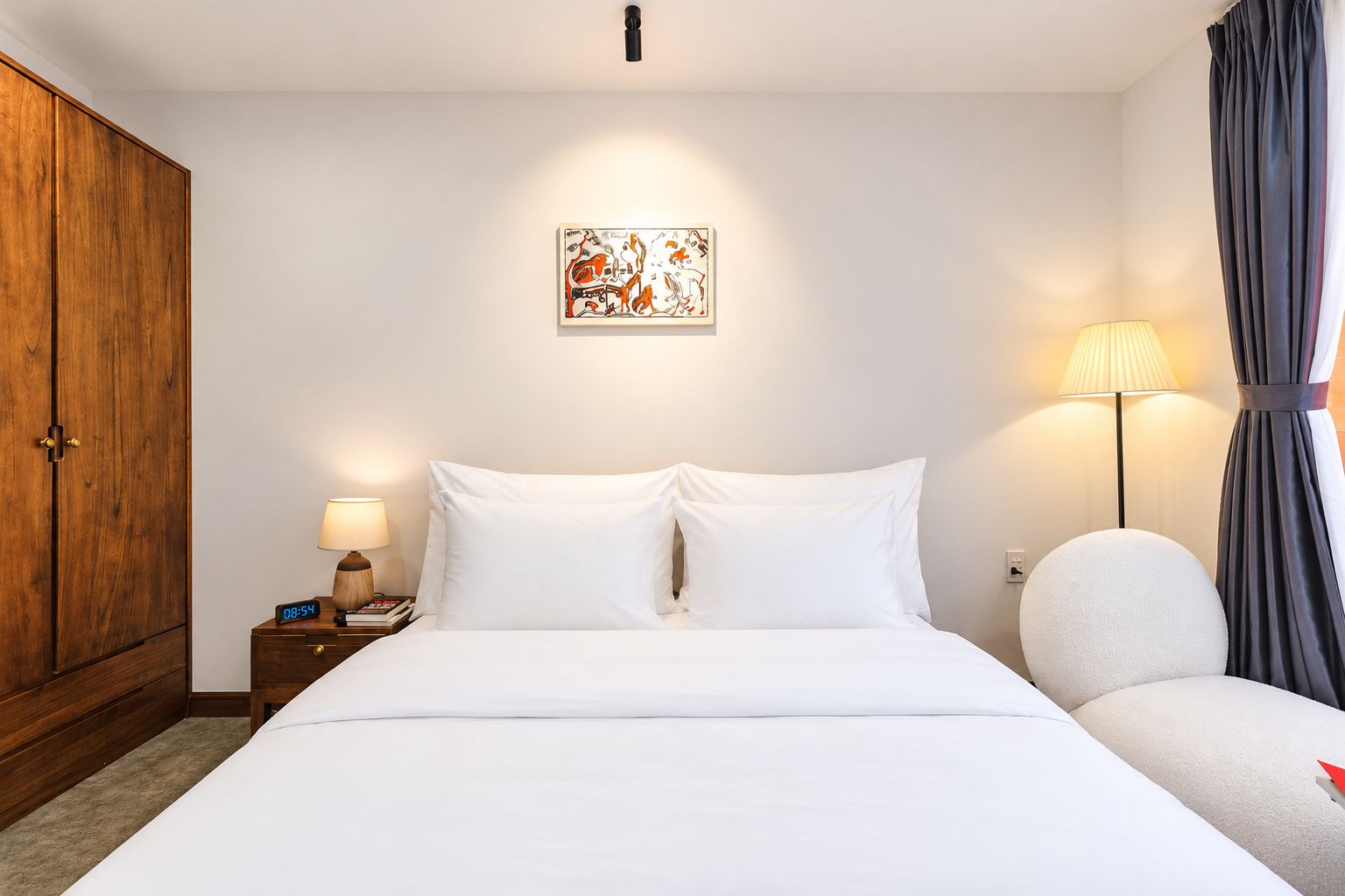 Une chambre d’hôtel avec un lit, une table de chevet avec une lampe, une armoire en bois, un tableau au mur et une chaise avec une lampe.