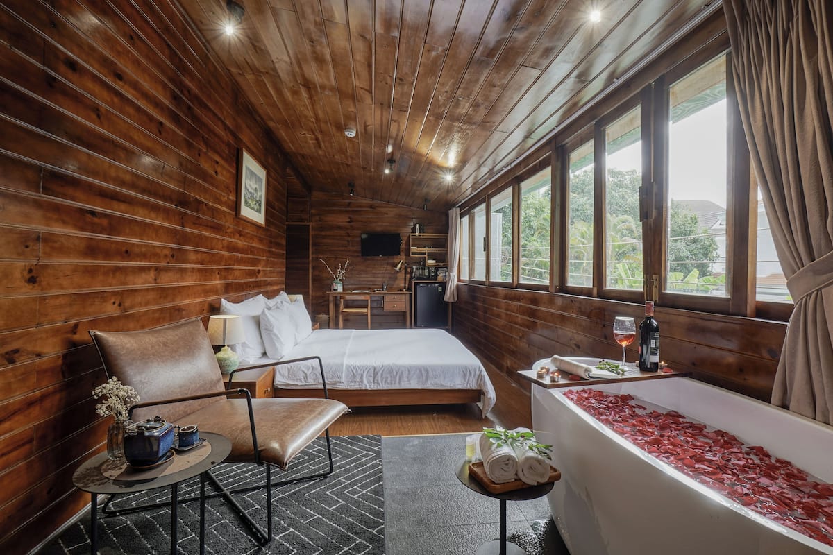 A wooden cabin room with a bed, a chair, a small table with a teapot, a bathtub with red bath mats, and a window with curtains.