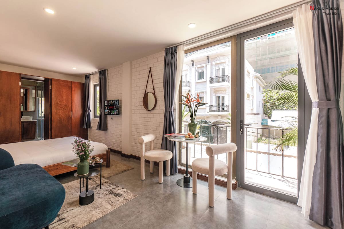 A modern hotel room with a bed, two chairs, a small table, and a balcony with a view of buildings and greenery.