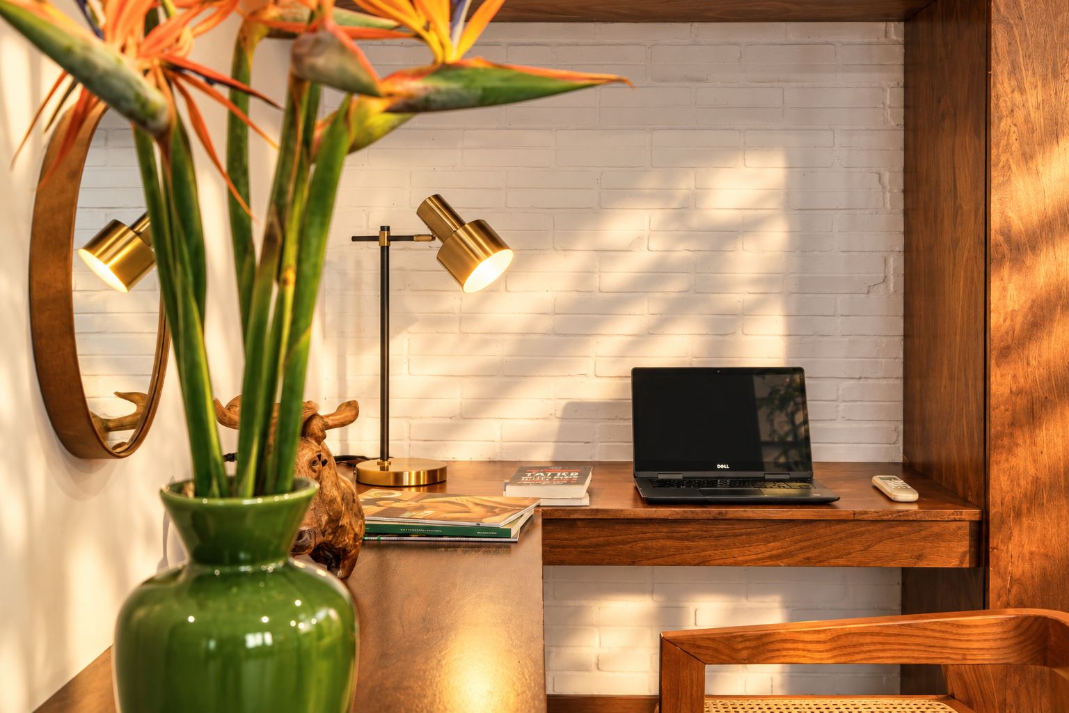 A wooden desk with a laptop, a mirror, a green vase with a plant, and two floor lamps.