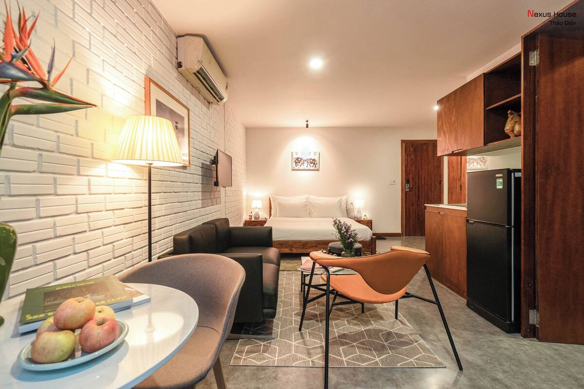 A modern hotel room with a bed, a small seating area with a table, a refrigerator, and a kitchen area with wooden cabinets.