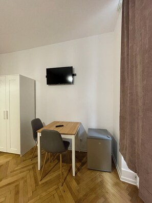 A small room with a wooden table, two chairs, a television mounted on the wall, a white wardrobe, and a small refrigerator.