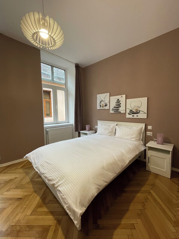 A bedroom with a large bed, a bedside table with a lamp, a window with curtains, and artwork on the wall.