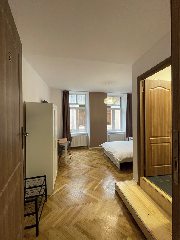 A compact hotel room with a bed, a desk, and a chair. There are windows with curtains, a radiator, and a wooden floor.