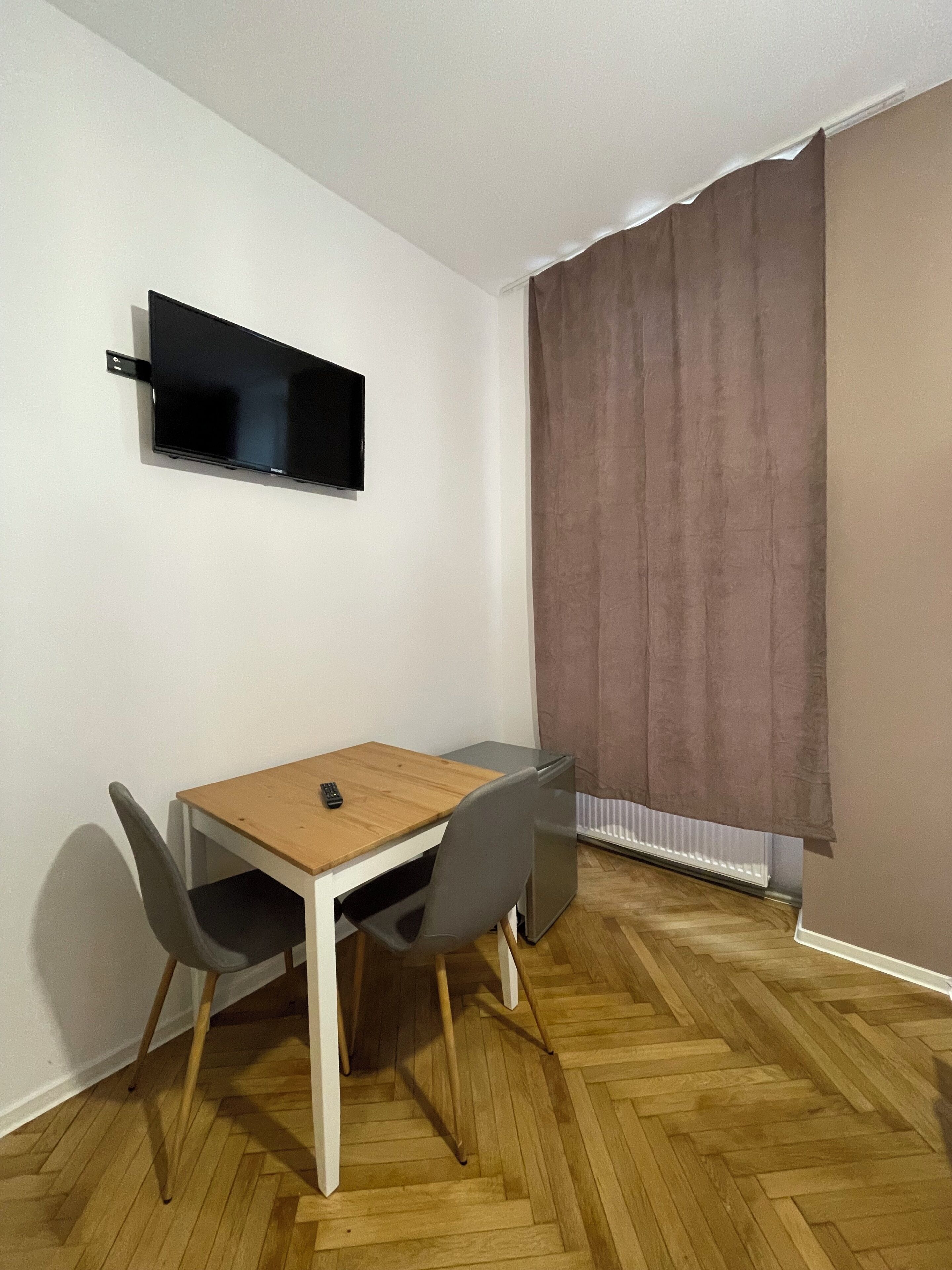A small, modern room with a wooden table and two chairs, a flat-screen TV mounted on the wall, and a window with curtains.
