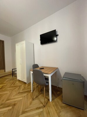 A small room with a wooden herringbone floor, a white wardrobe, a desk with a chair, a flat-screen TV mounted on the wall, and a small refrigerator.