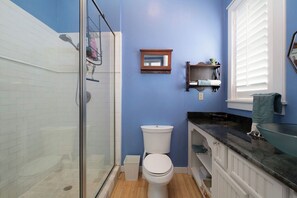 A bathroom with a glass shower enclosure, a white toilet, a blue wall, and a black countertop. - Bridges Cottage - Vintage in Heart of Cedar Key! (Cedar Key)