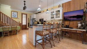 A modern kitchen with wooden cabinets, a central island with bar stools, a dining area with a table and chairs, and a large TV mounted on the wall. - Bridges Cottage - Vintage in Heart of Cedar Key! (Cedar Key)