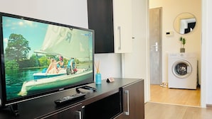 Una sala de estar moderna con un televisor de pantalla plana, un mueble de madera oscura, una lavadora y una pequeña planta.