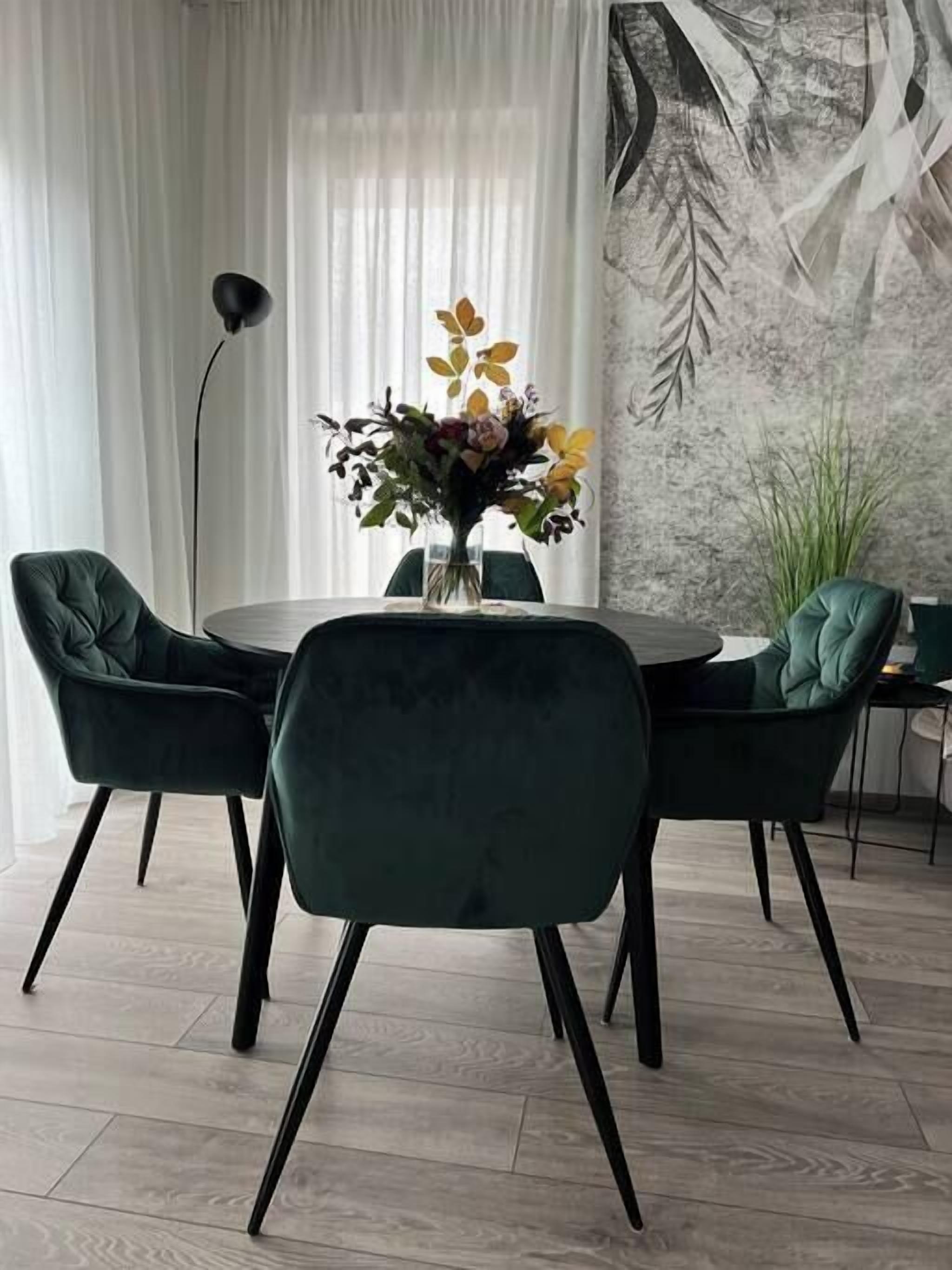 Un comedor con una mesa redonda, cuatro sillas tapizadas de color verde, un jarrón con flores y una pared con un diseño de hojas.