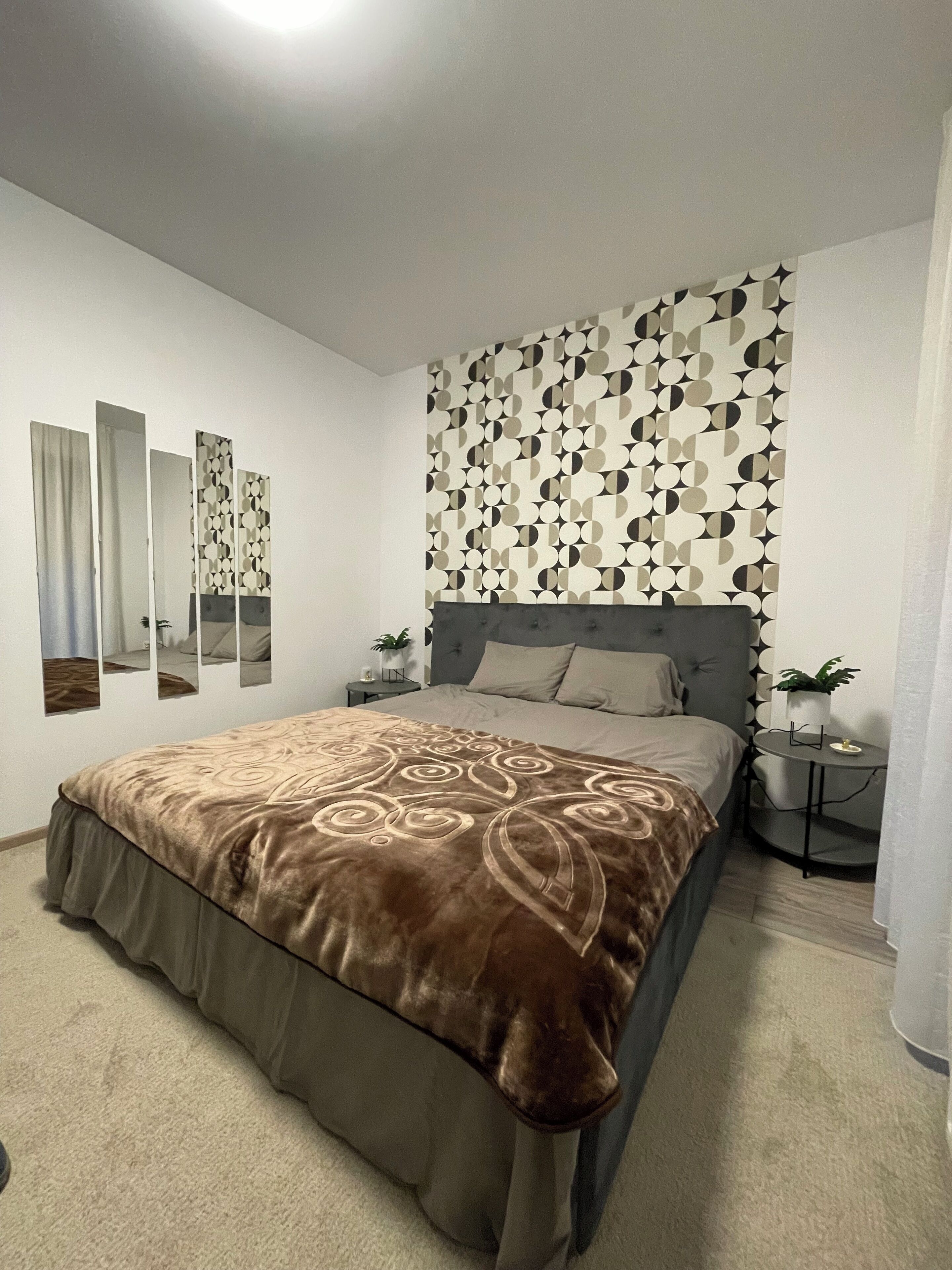 Un dormitorio con una cama grande, cabecera con diseño geométrico, mesita de noche con una planta y pared con motivo circular.