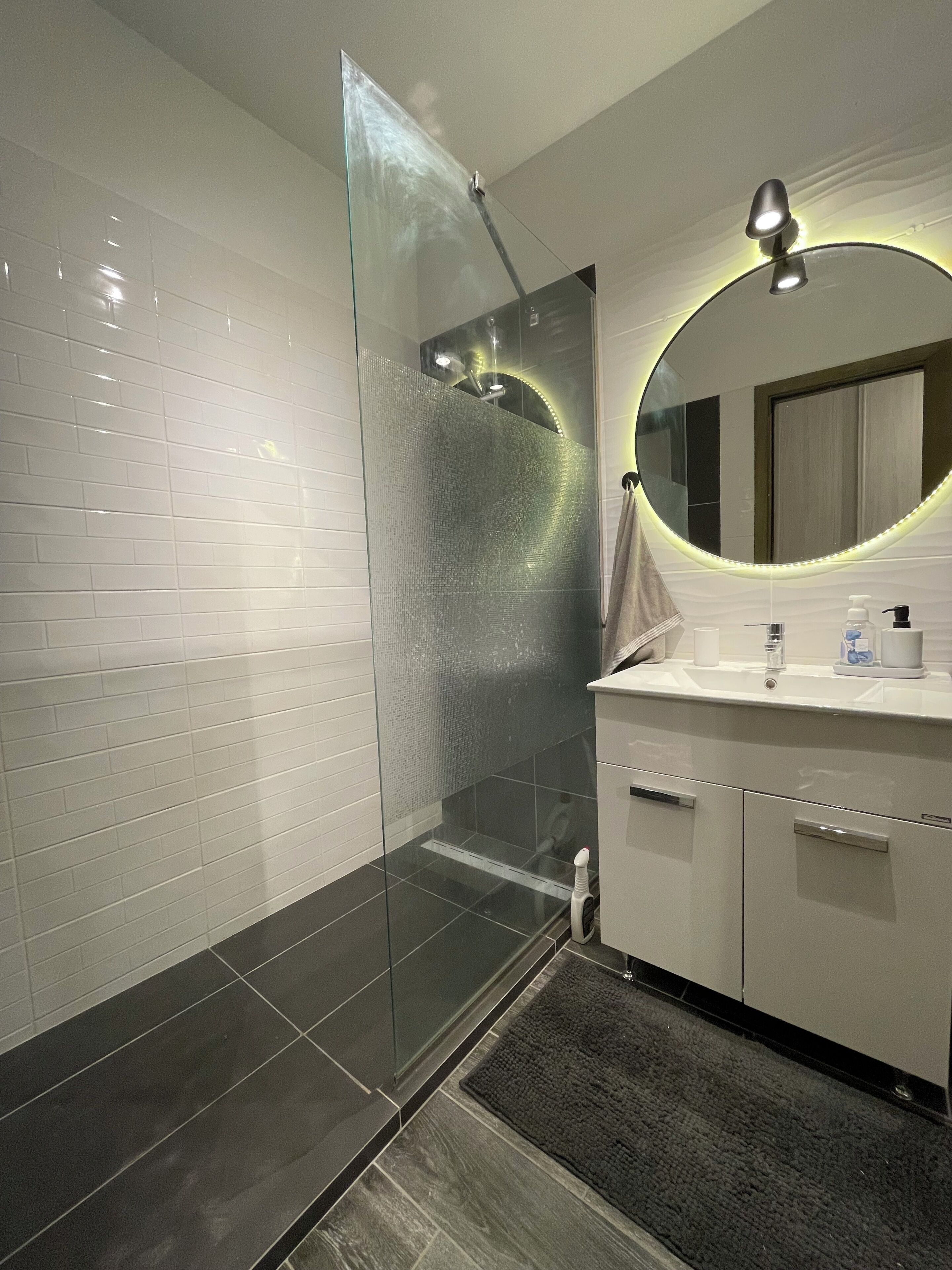 Un baño moderno con cabina de ducha de vidrio, espejo redondo con luces LED y tocador blanco con lavabo y dispensador de jabón.
