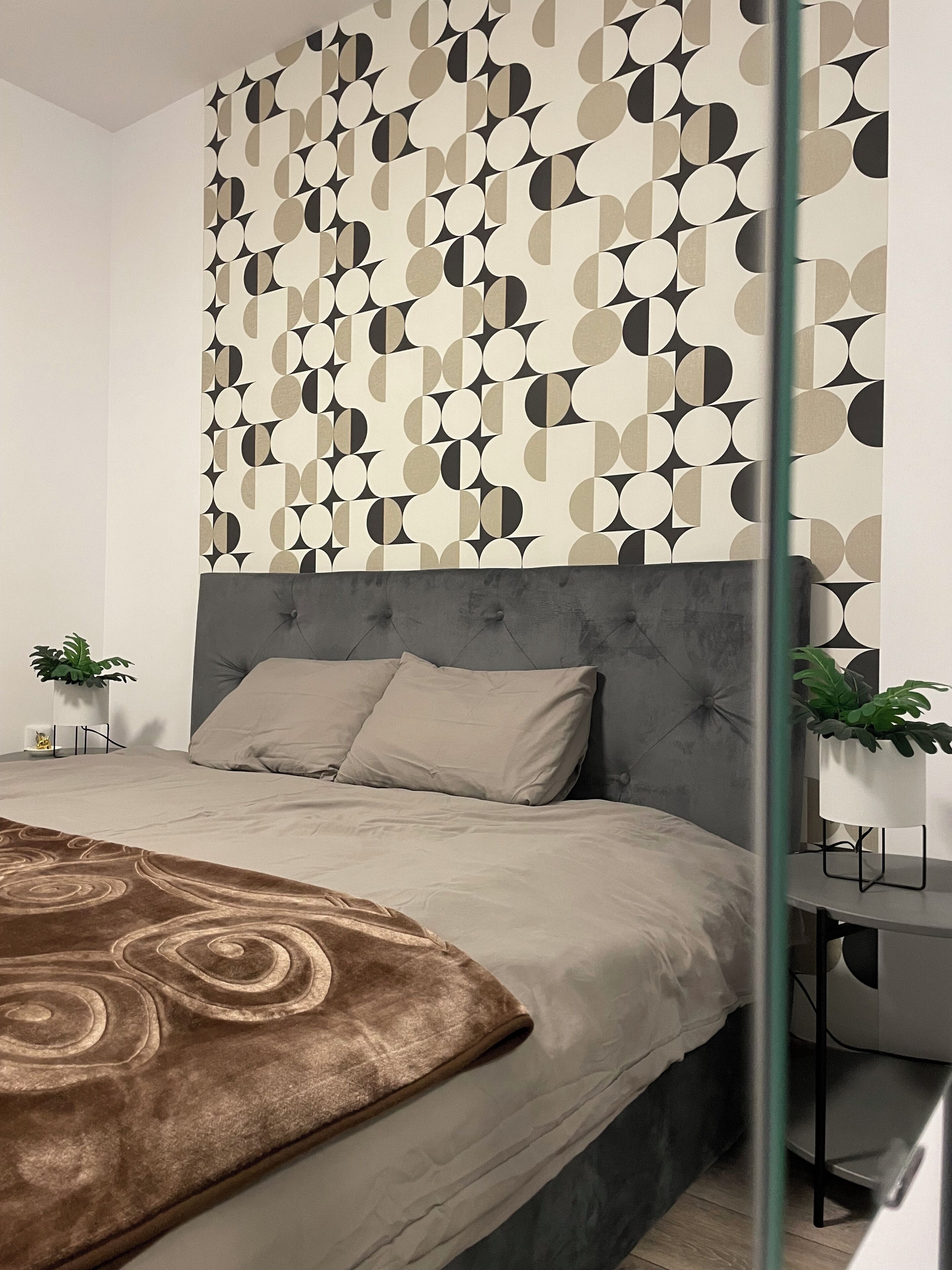 A modern bedroom with a geometric-patterned wall, a tufted headboard, a brown throw blanket, and two potted plants.