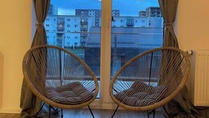 A room with two wicker chairs facing a sliding glass door with curtains, wooden flooring, and a view of buildings.